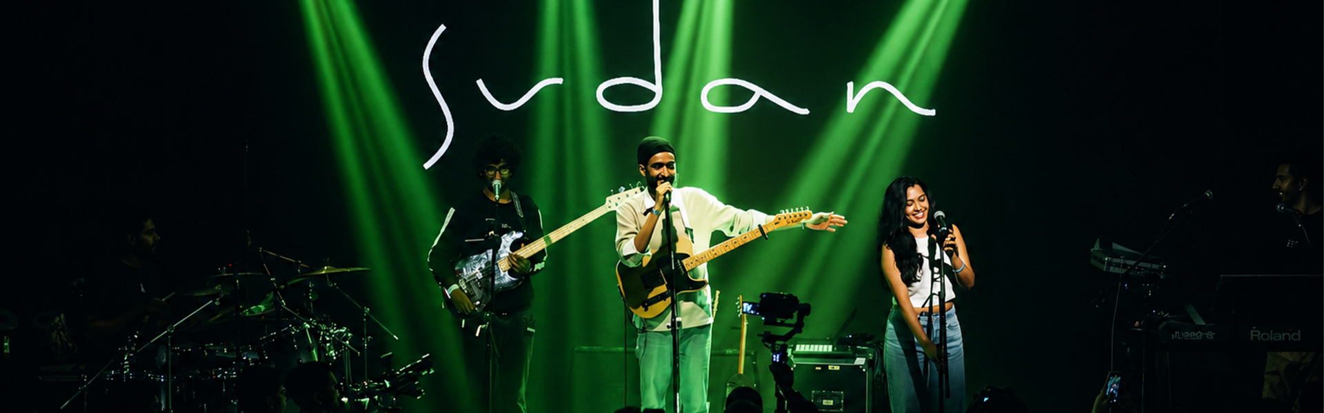 Sudan performing live on stage in Mumbai with his band under green lighting, with the Sudan logo projected behind him.